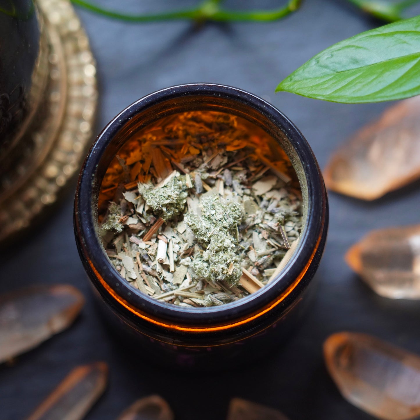 Mugwort and Herbs stored in amber jar, surrounded by orange crystals, a house plant and meditation tool in the background