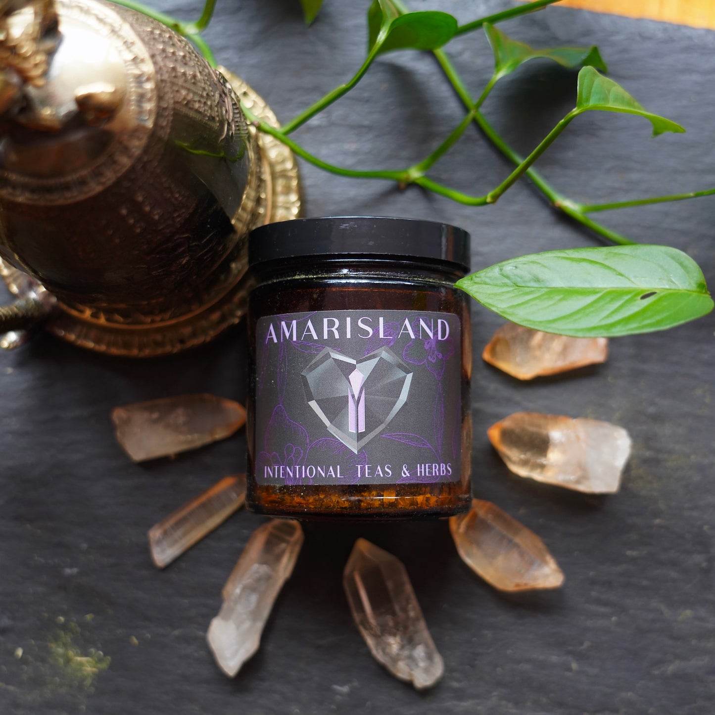 Jar of Tea and Herbs surrounded by hematite quartz crystals, on a slate slab with a house plant and meditation bell in the background