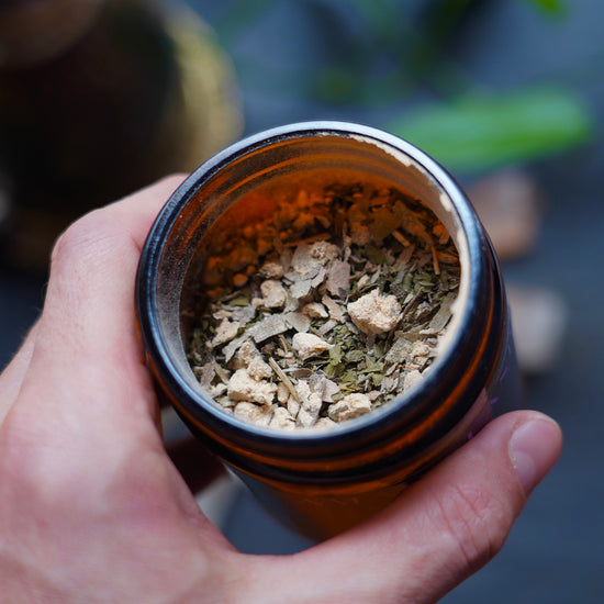 Powder of herbs and leaves of tea in an amber glass jar, held in the left hand over a black slate and meditation tools in the background